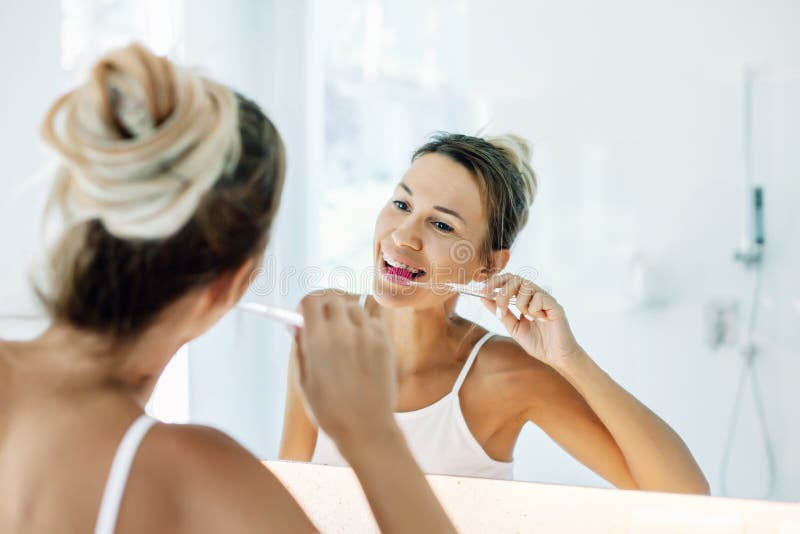 Photo of woman looking at mirror, holding toothbrush and brushing teeth. Making everyday morning routine in bathroom. Adult human with dental braces doing daily oral care. Photo of woman looking at mirror, holding toothbrush and brushing teeth. Making everyday morning routine in bathroom. Adult human with dental braces doing daily oral care